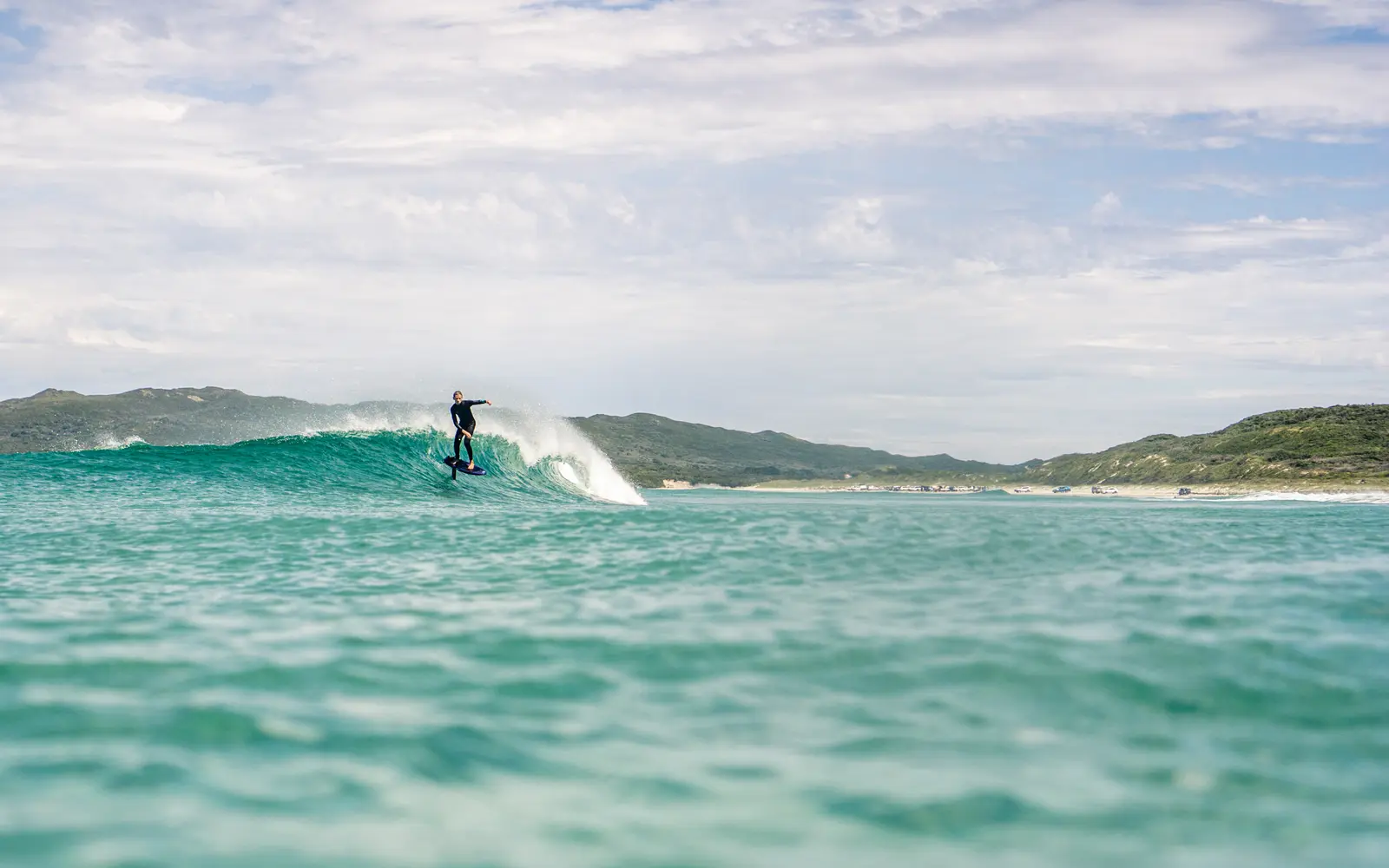 A prone foiler carves a turquoise wave, with a hilly coastline in the background