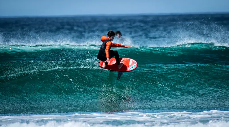 A prone foiler in an orange wetsuit rides a red board above a clean teal wave