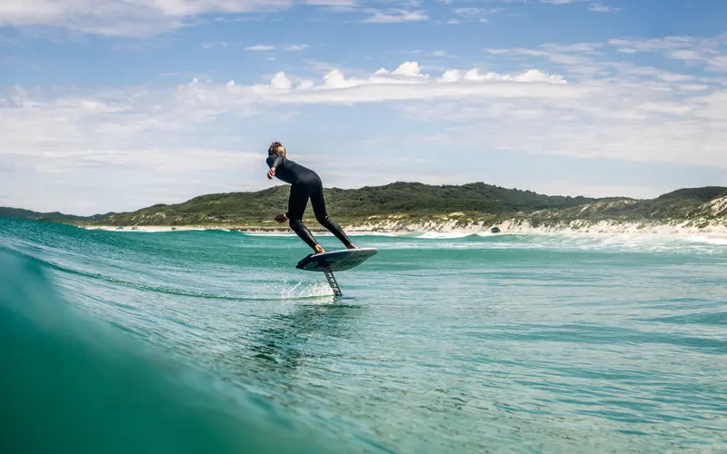 A prone foiler glides across a wave, viewed from behind