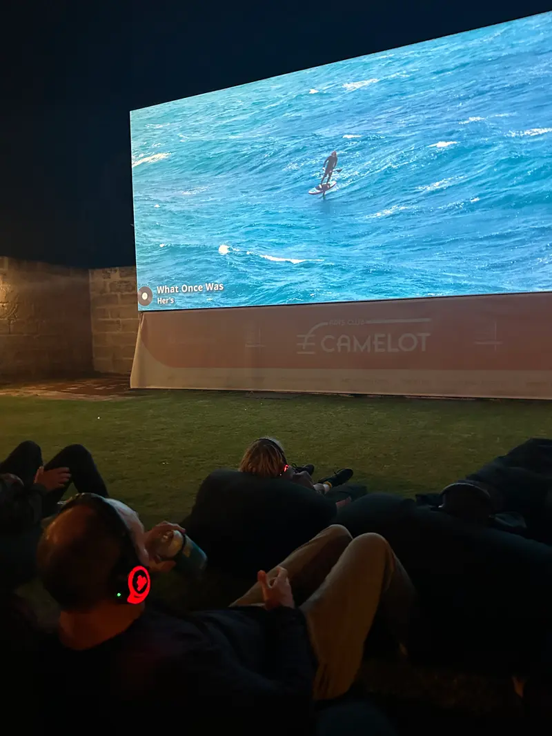 A crowd lies on the grass at the outdoor cinema, watching downwind foiling footage light up the screen