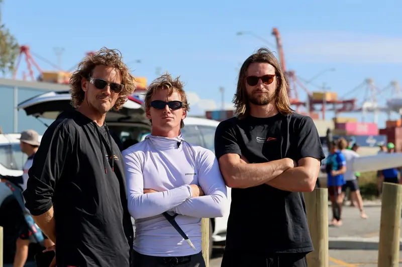 Three crew members stand together at the event site, port cranes in the background