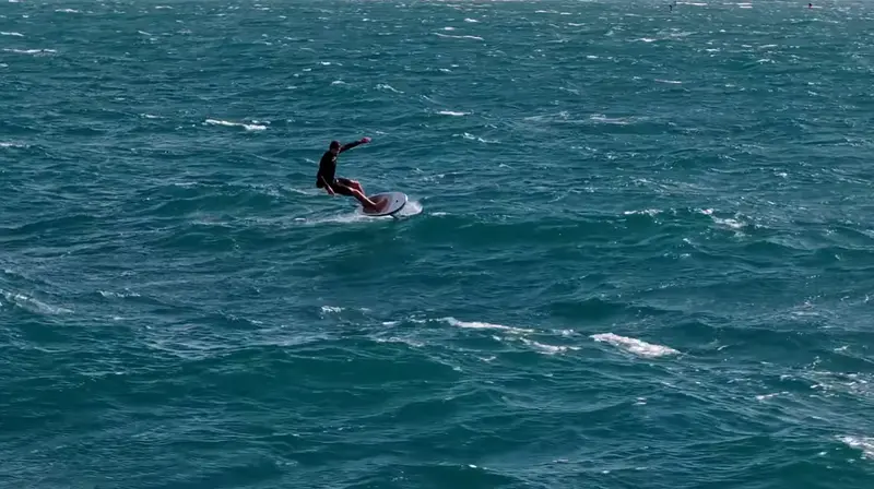 A downwind foiler rides across open teal water, seen from above