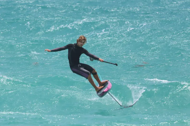 A downwind SUP foiler mid-carve on a pink board, holding a paddle, above vivid turquoise water