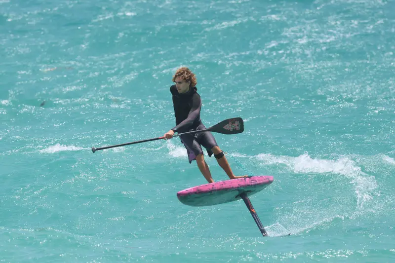 A foiler carves along a wave face on a pink downwind board, paddle blade skimming the water