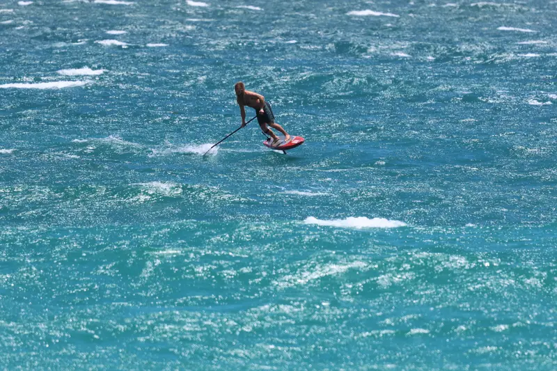 A foiler rides a red downwind board across open blue water, paddle in hand