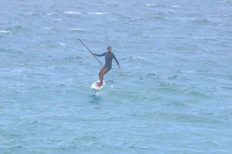 A foiler in a wetsuit leans into a turn on a white downwind board, paddle held out wide