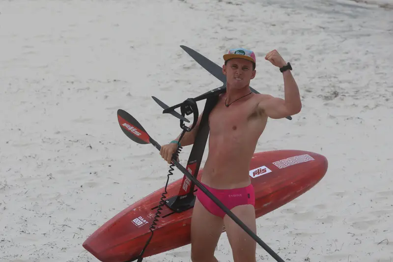 A rider flexes on the sand next to a red board and foil