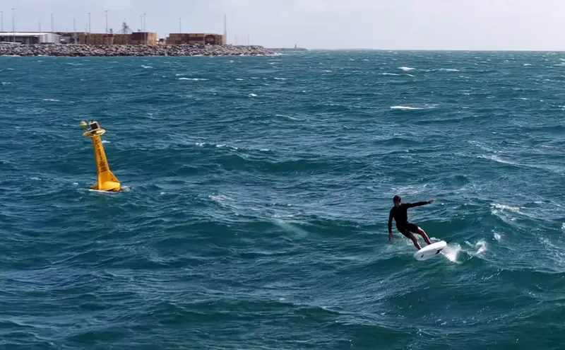 A downwind foiler glides past a channel marker, with a working harbour in the distance